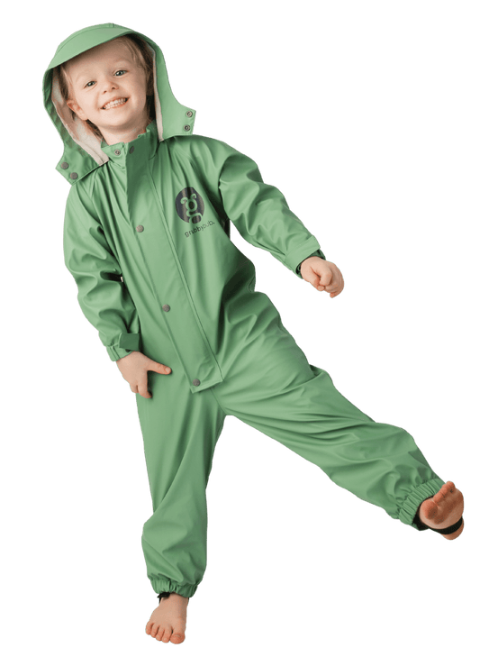 Happy girl in a sage green waterproof toddler puddle suit standing on one foot