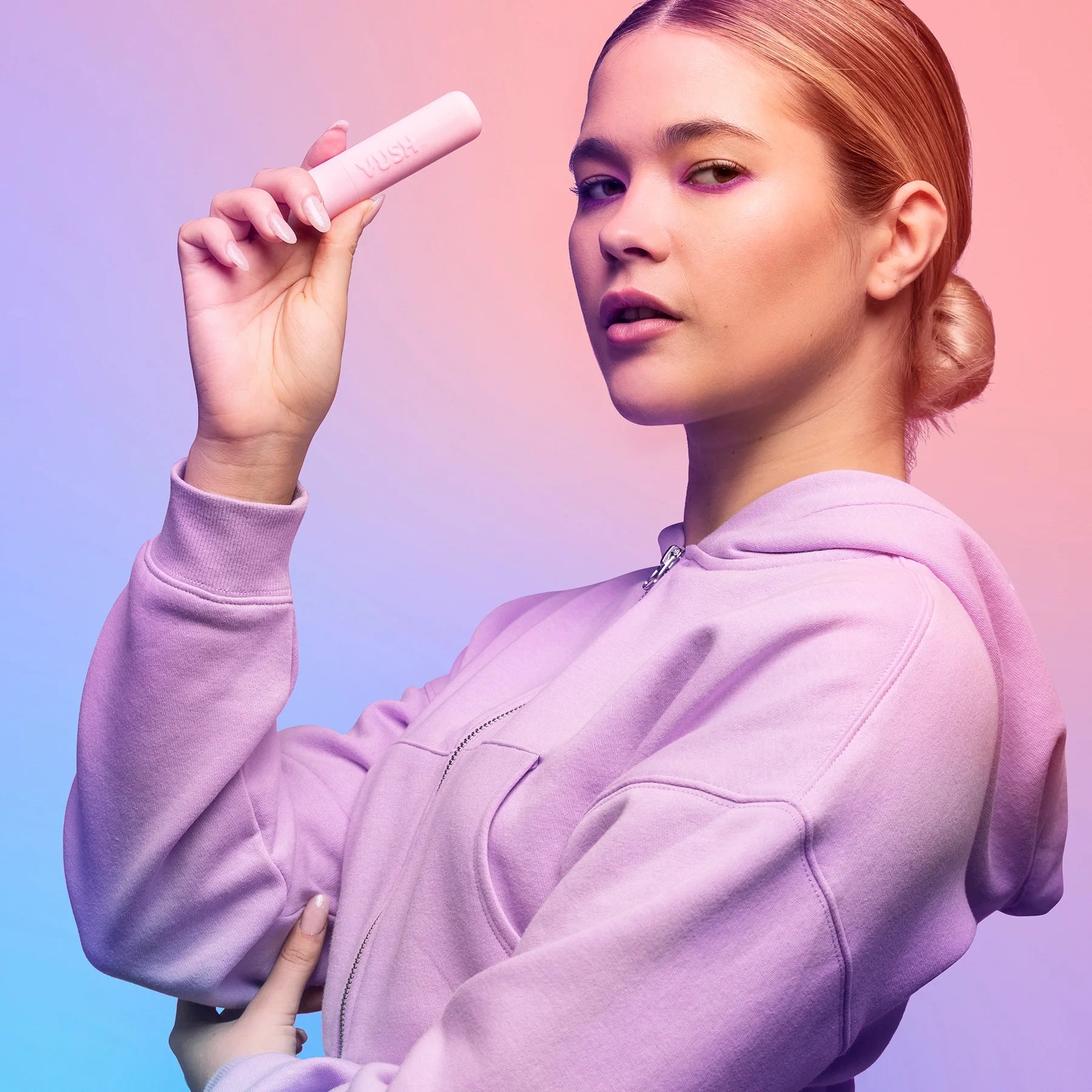 Woman with blonde hair in low bun wearing purple hoodie, holding pink VUSH Gloss Bullet Vibrator in case.