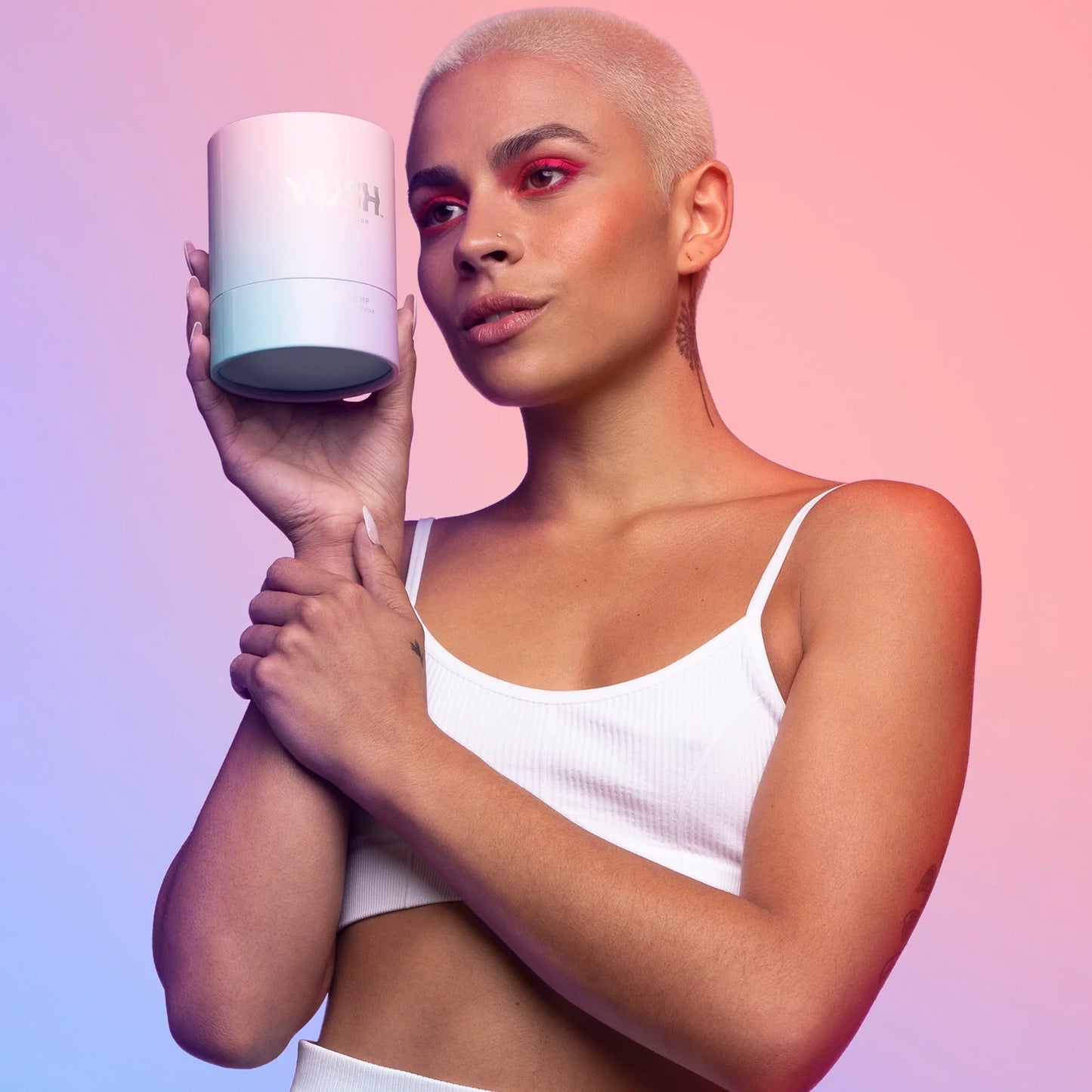 Person with bleached shaved hair wearing white crop top posing with pink/purple/blue cylinder packaging in right hand, holding right arm with left hand.